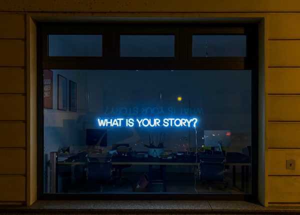 Ein Fenster, das den Blick in ein dunkles Büro mit Schreibtisch freigibt. Im Fenster die Leuchtschrift: What is your Story?