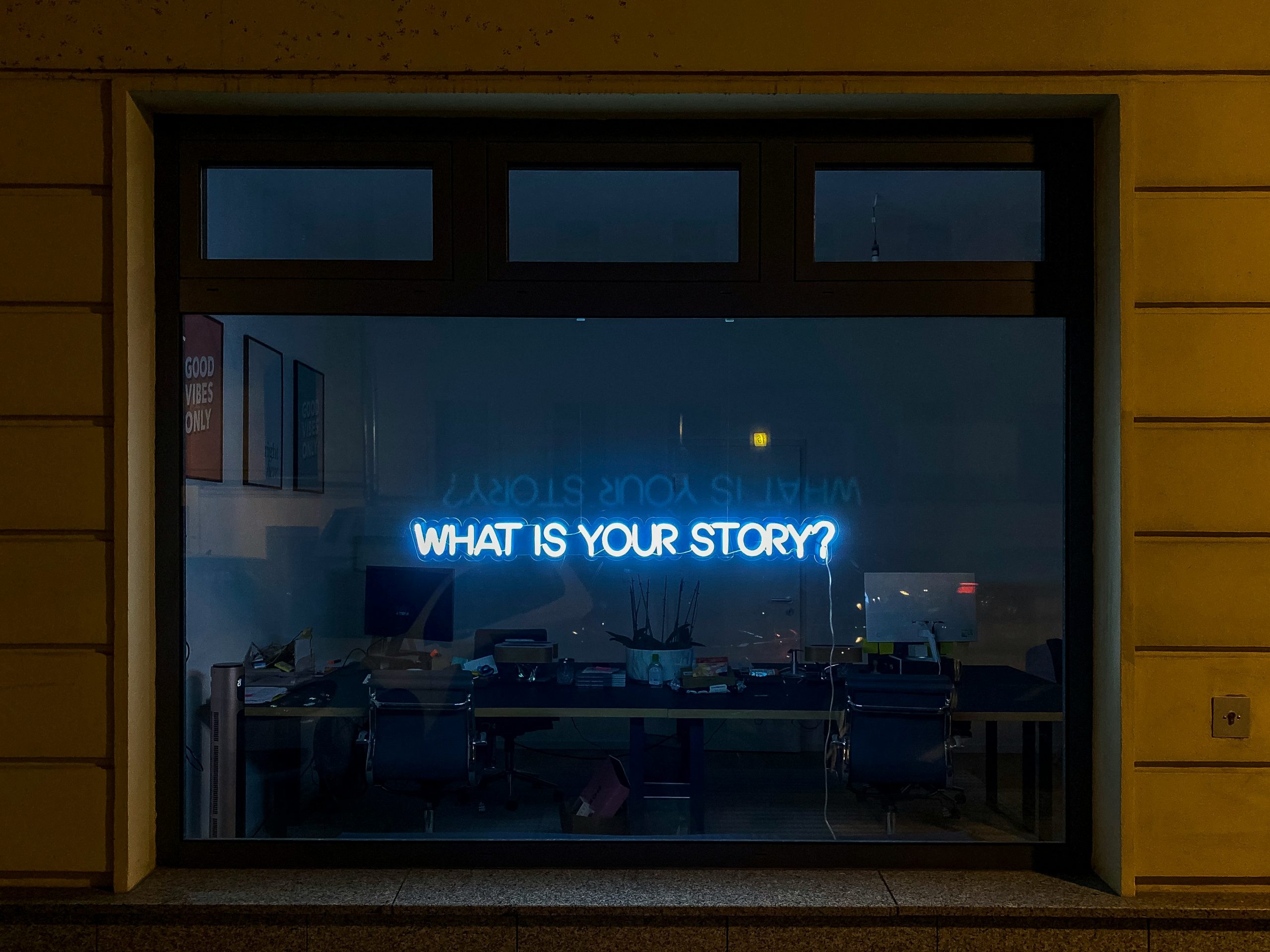Ein Fenster, das den Blick in ein dunkles Büro mit Schreibtisch freigibt. Im Fenster die Leuchtschrift: What is your Story?