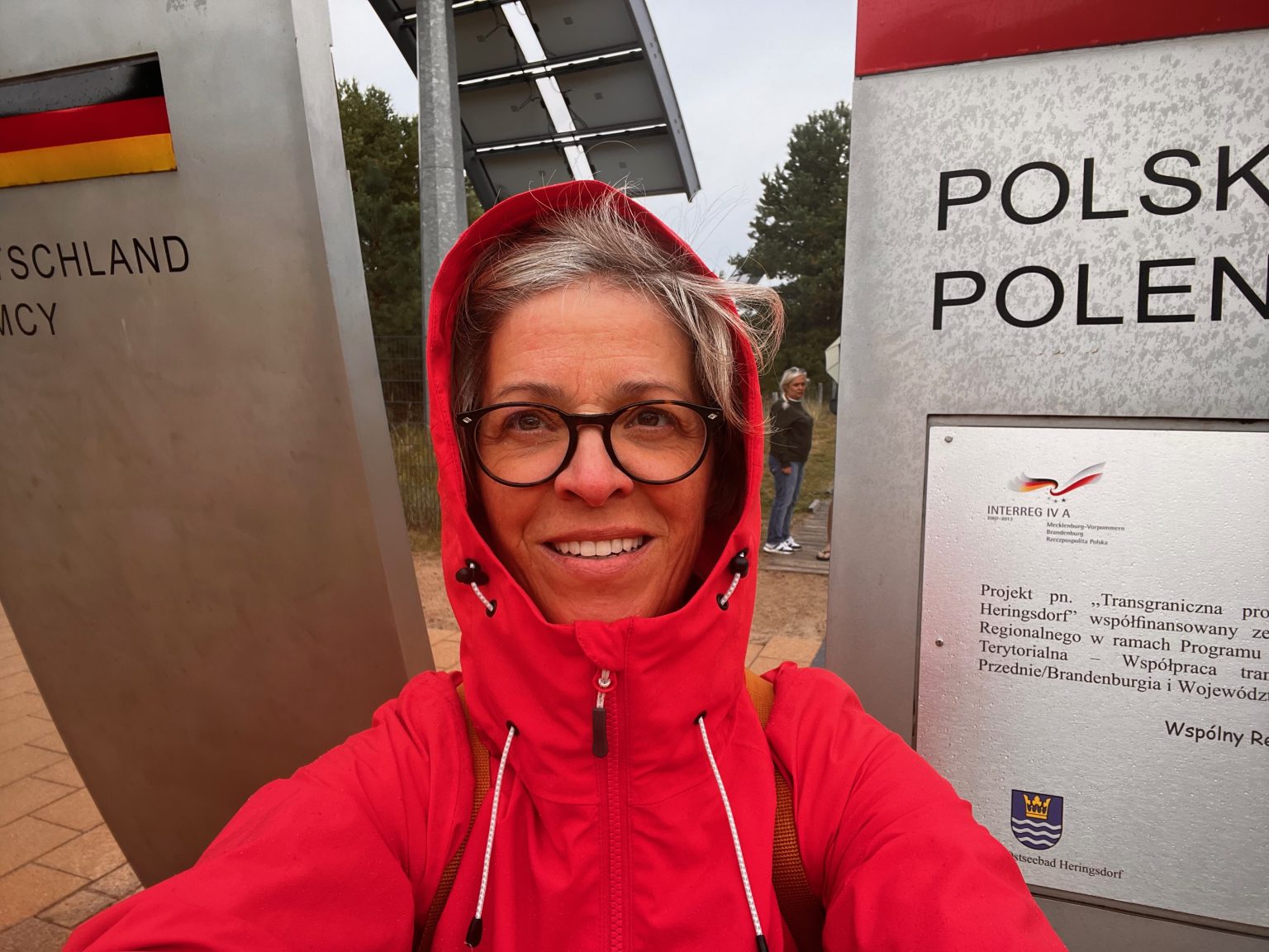 Eine Frau in Regenjacke bei starkem Wind vor den beiden Grenzschildern an der deutsch-polnischen Grenze auf Usedom.