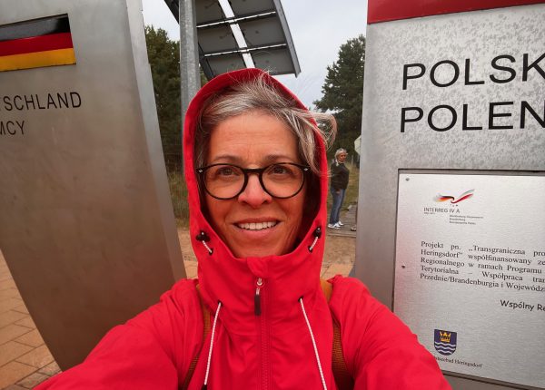 Eine Frau in Regenjacke bei starkem Wind vor den beiden Grenzschildern an der deutsch-polnischen Grenze auf Usedom.